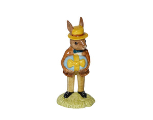 The bunny is wearing a brown jacket, blue bow tie and a yellow hat with a red band. He is holding a large easter egg, wrapped in blue with a big yellow bow.